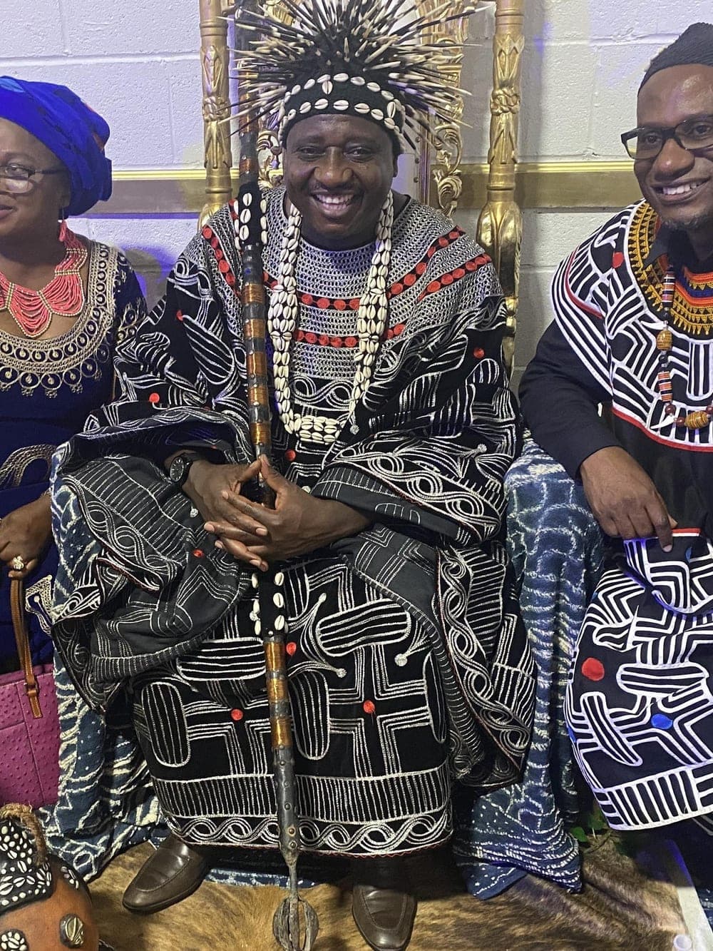 HRH Fon Fomuki Walters Ticha IX seated on the throne in full Guneku regalia — a feathered and cowrie headdress, an embroidered gown and a beaded staff — with two guests either side.
